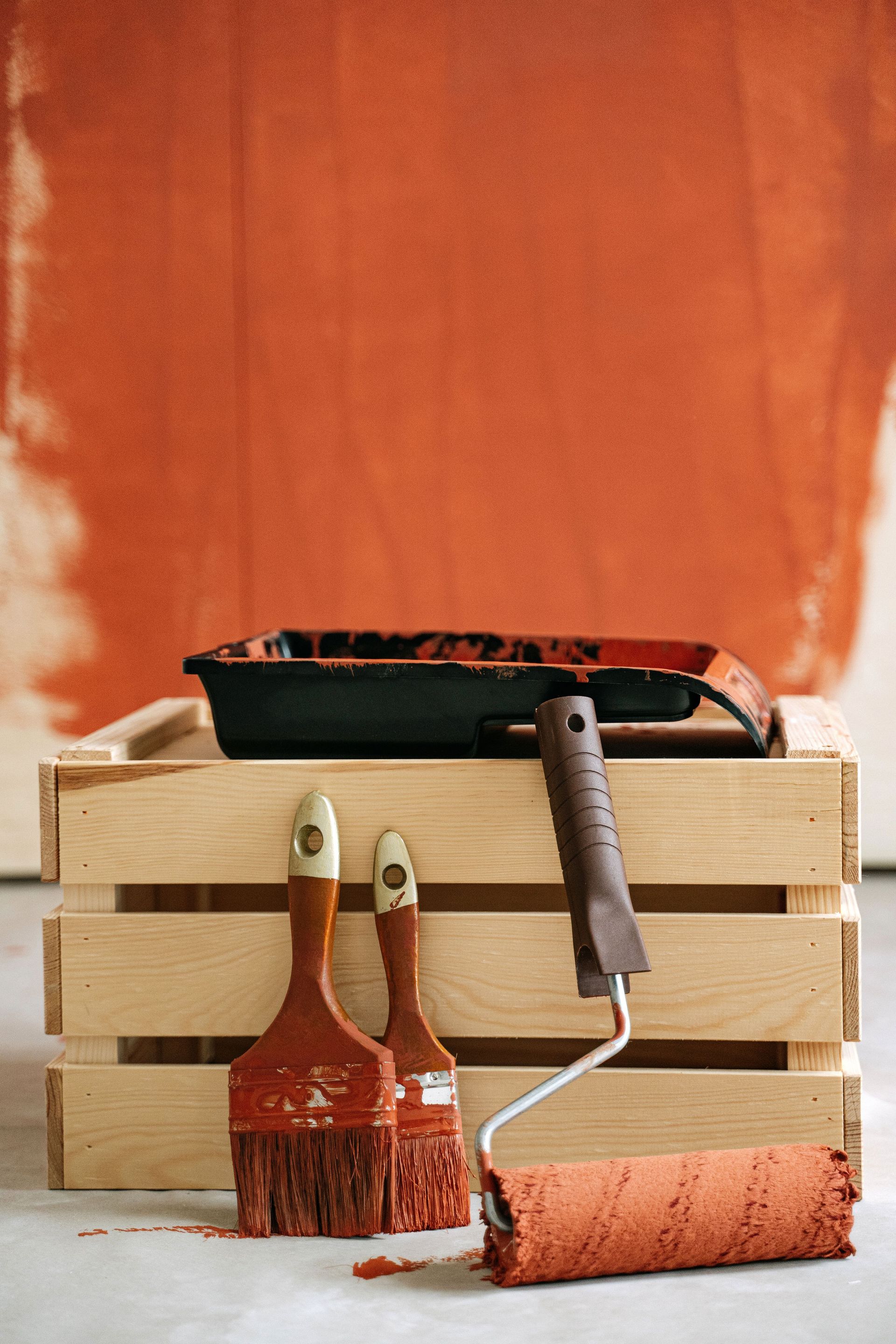 Wooden crate with paintbrushes and roller against a partially painted wall.